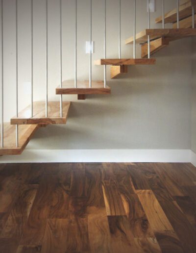 Modern floating wooden staircase against a white wall in a room with hardwood flooring.